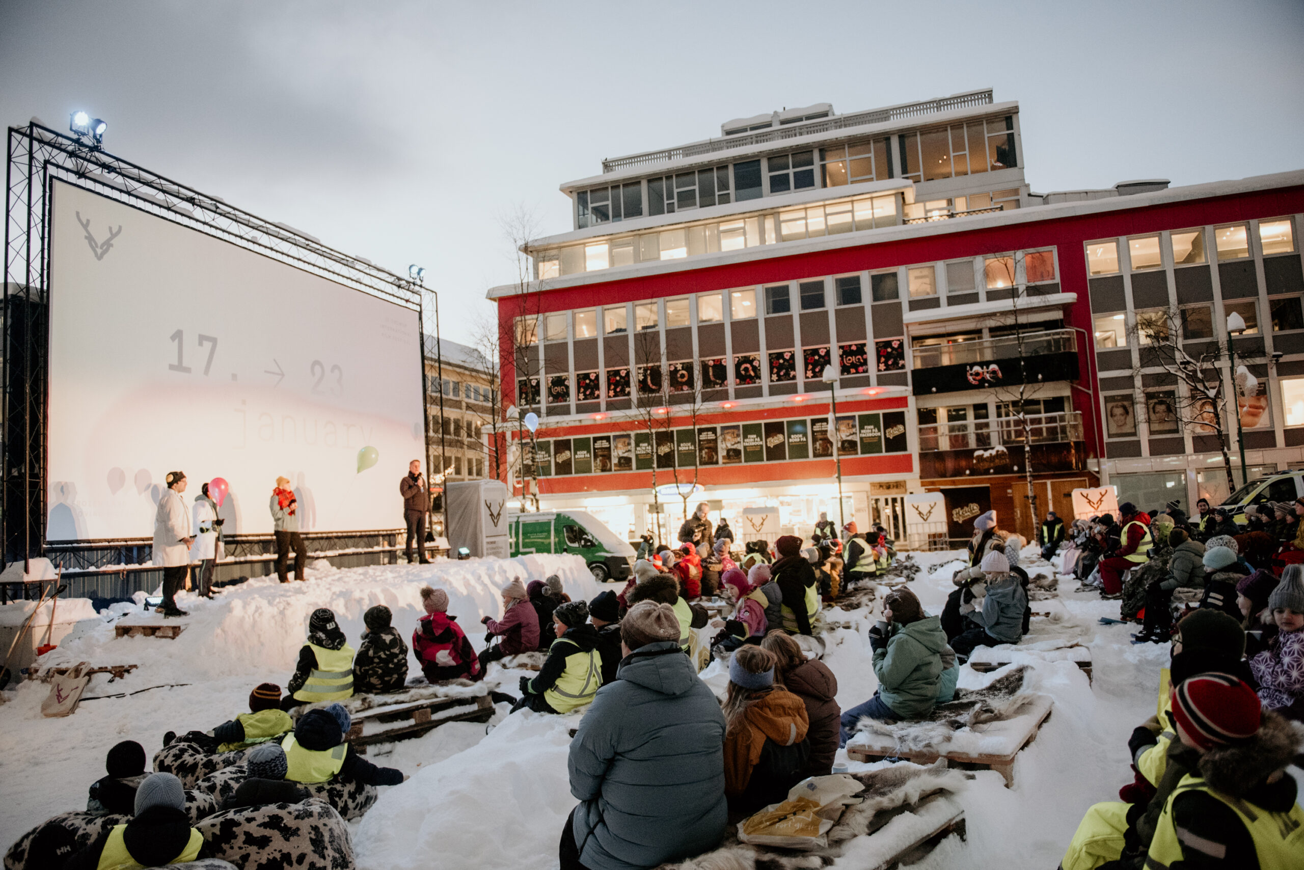 Barn og voksne sitter utendørs i snø foran et stort filmlerret i Tromsø