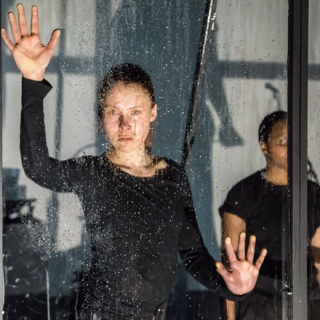 Ung kvinne kledd i svart ser i kamera gjennom et stort vindu med vanndråper på. Holder håndflatene mot vinduet.